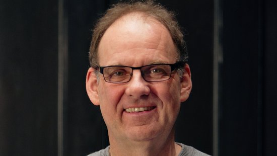 Das Bild zeigt einen älteren Forscher im grauen Pulli mit dunkel umrandeter Brille vor einer dunkel vertäfelten Wand in die Kamera lächeln.