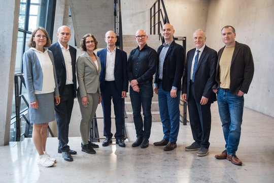 A group of eight people, five men and three women, stand in a line indoors against a concrete wall. They are dressed in business and casual wear, exuding a professional yet relaxed atmosphere.