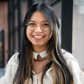 Mia Le Bild von Mia Le - Sie trägt eine naturfarbene Jacke über einem roséfarbenen Pullover mit weißem Kragen. Sie hat dunkle, lange Haare, eine Brille und steht vor einer Fensterfront.