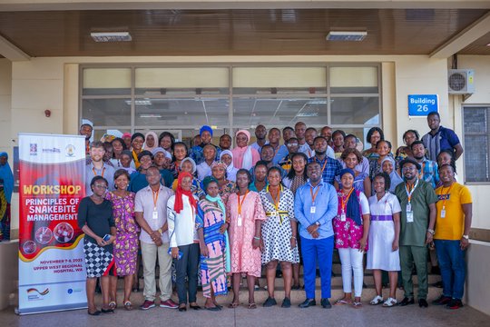 The picture shows a group of people standing in front of a hospital entrance next to a banner saying 'Workshop - Principles of Snakebite Management'. Everyone is looking friendly into the camera.