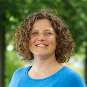 Dr. Anke Thielebein: eine Forscherin mit lockigen, halblangen Haaren in einem leuchtend blauen Pullover. Sie steht vor einem unscharfen Blätterhintergrund.