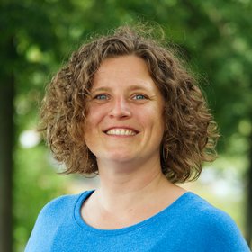 Dr. Anke Thielebein Dr. Anke Thielebein: eine Forscherin mit lockigen, halblangen Haaren in einem leuchtend blauen Pullover. Sie steht vor einem unscharfen Blätterhintergrund.