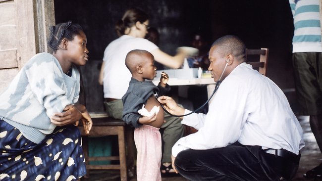 Ein ghanaischer Arzt hockt vor einem Kind, dahinter dessen Mutter. Der Arzt untersucht das Kind mit einem Stetoskop. Im Hintergrund arbeitet eine Kollegin des BNITM Hamburg mit dunklen Haaren an einem Tisch.