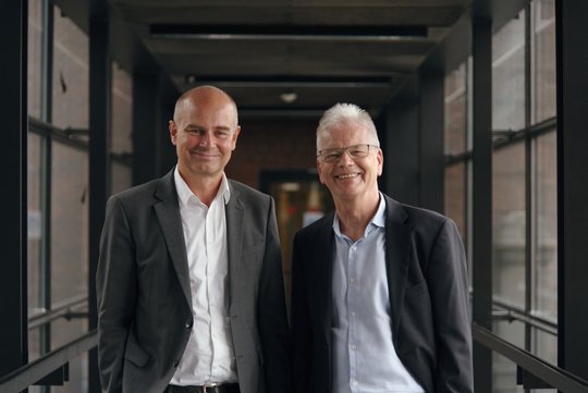 The new and the old CEO stand in the transition between the old and the new building. Both are wearing a white shirt and a dark jacket.
