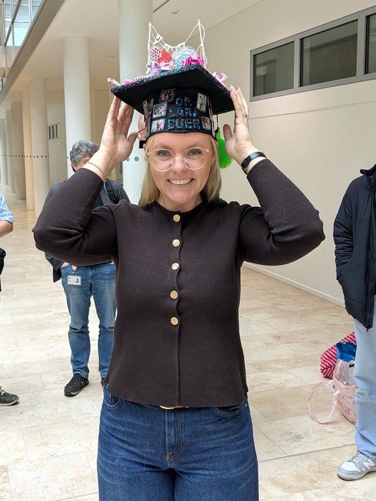 The photo shows a close-up of Lara Buer after her exam. She is proudly holding her doctoral hat with both hands and smiling at the camera. The black cap, adorned with many colorful details, was handmade by her colleagues in Borstel. Lara is wearing a chic brown cardigan with gold buttons.
