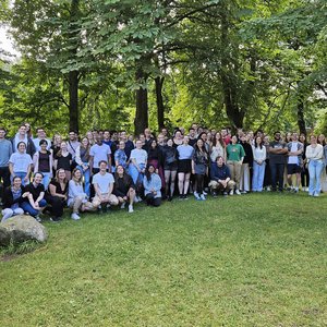 Großes Gruppenfoto von etwa 70 Personen auf einer grünen Wiese unter Bäumen. Die Gruppe besteht aus jungen Erwachsenen, die locker zusammenstehen und in die Kamera lächeln. Es ist sonnig und sommerlich.