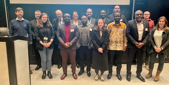 Das Bild zeigt eine diverse Gruppe an Forscherinnen und Forschern, die vor einer Leinwand stehen und freundlich in die Kamera lächeln.