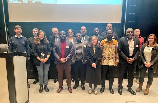 The picture shows a group of researchers from Europe and Africa standing in front of a screen and smiling friendly into the camera.