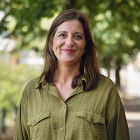 Dr Sophie Duraffour: a woman in a green blouse and black trousers on a sunny path with trees in the background. She has dark, long hair.