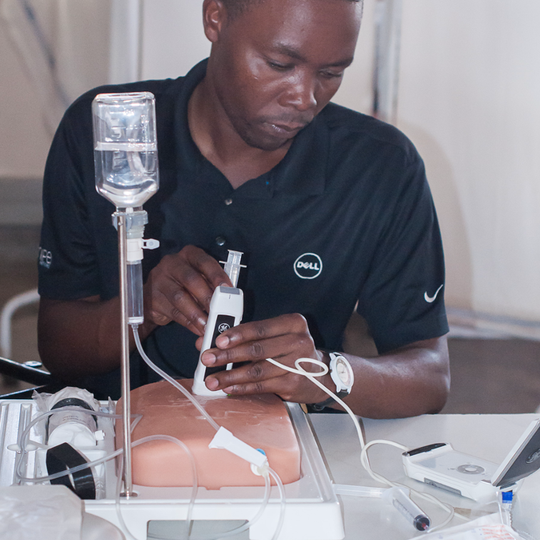 Das Bild zeigt einen jungen Mann, der ein kleines Ultraschallgerät mit einem Übungsmaterial benutzt. Das Ultraschallgerät befindet sich auf dem Übungsmaterial und er schaut auf einen kleinen Bildschirm, auf dem das Ultraschallbild angezeigt wird.