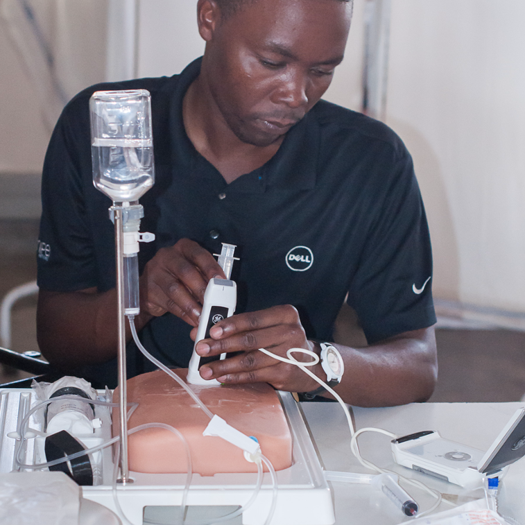 The picture shows a young man using a small ultrasound machine with an exercise material. The ultrasound machine is on the exercise material and he is looking at a small screen displaying the ultrasound image.