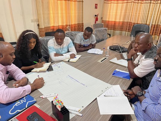 Sechs Personen sitzen mit Notizbüchern und Papieren um einen Konferenztisch und diskutieren. Auf dem Tisch liegen Stifte, ein Laptop und ein großes Blatt Papier mit Diagrammen.