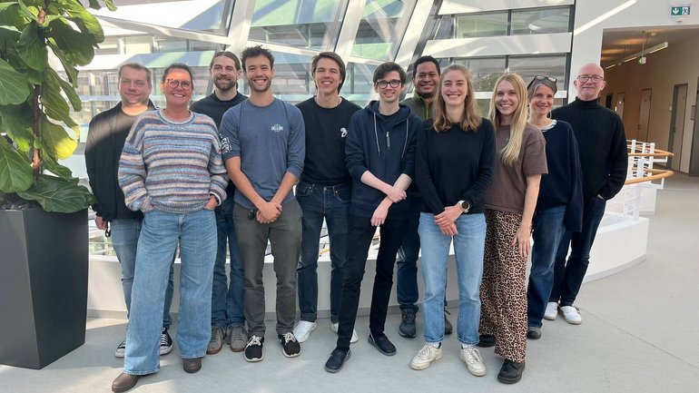 Gruppe von elf Personen steht in einem hellen, modernen Innenraum mit Glasdach und Pflanzen, alle lächelnd und in legerer Kleidung