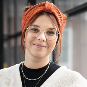Foto von Carolina van Gelder. Sie trägt einen naturfarbenen Pullover über einem dunklem Top und ein orangefarbenes Haarband und eine Brille. Sie steht vor einer Fensterfront., steht