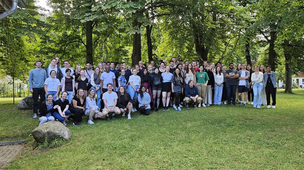 Großes Gruppenfoto von etwa 70 Personen auf einer grünen Wiese unter Bäumen. Die Gruppe besteht aus jungen Erwachsenen, die locker zusammenstehen und in die Kamera lächeln. Es ist sonnig und sommerlich.