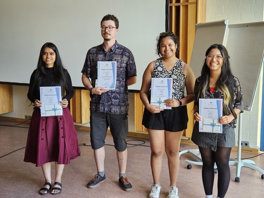 Vier junge Erwachsene stehen nebeneinander in einem Seminarraum und halten Zertifikate in der Hand. Sie lächeln in die Kamera. Im Hintergrund sind eine Leinwand, Flipcharts und Holzelemente sichtbar.