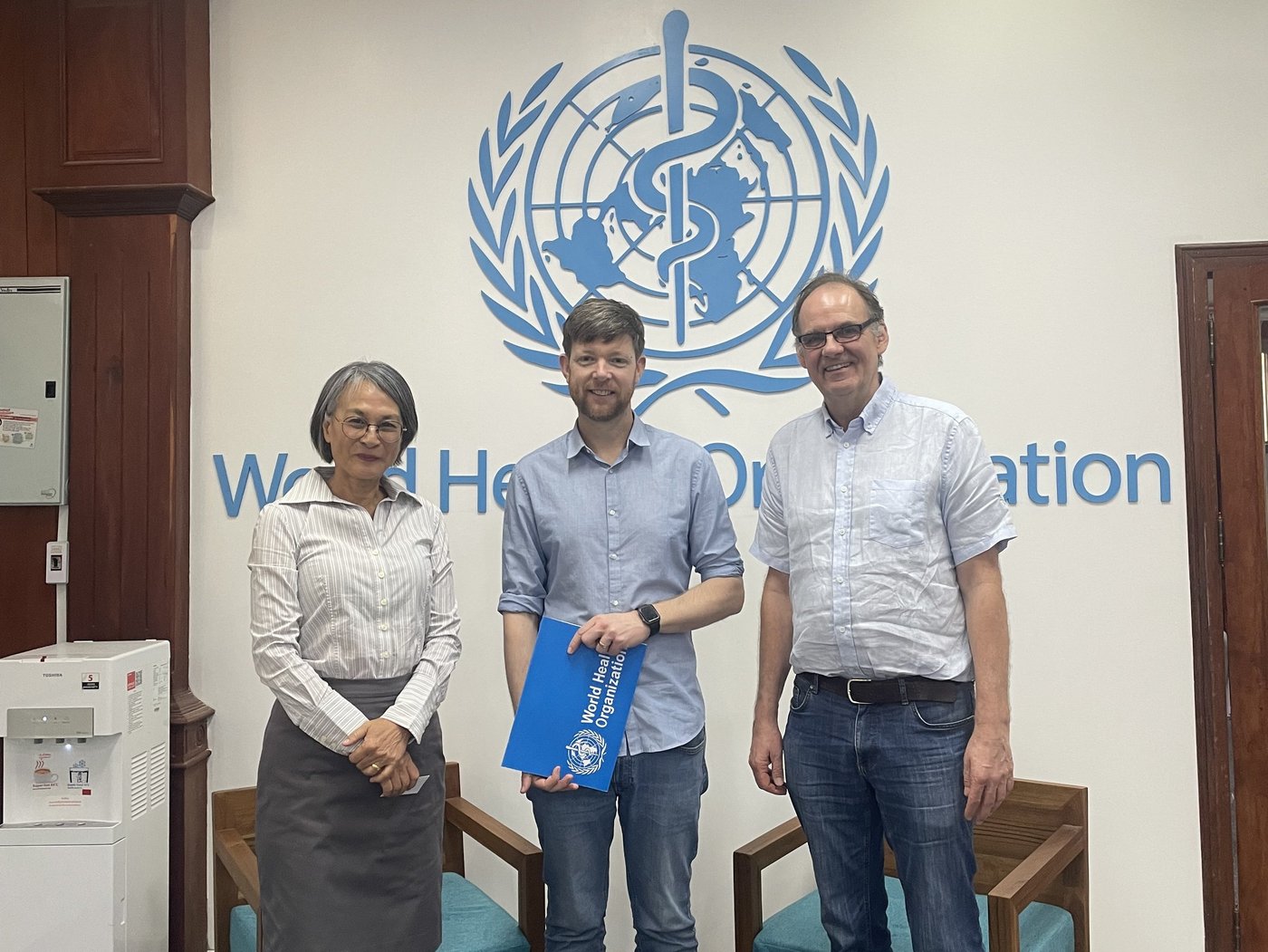 The picture shows three people standing in front of a WHO sign, a woman on the left and two men in the middle and on the right. They are all smiling at the camera. The man in the middle is holding a WHO folder.