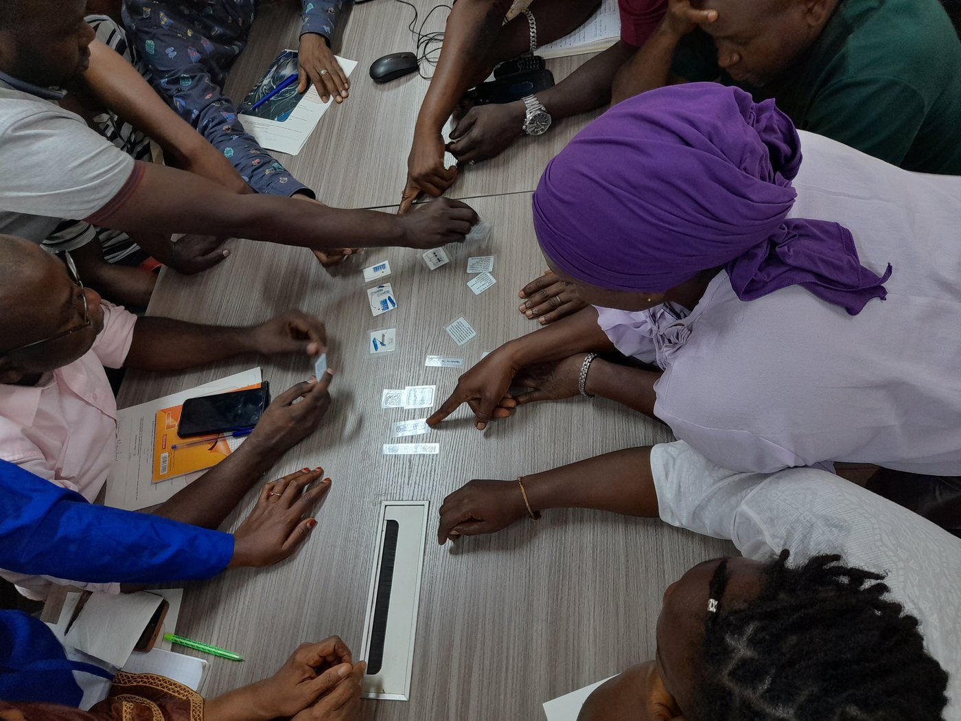 Lab staff from CRV-LFHVG performing a group working task all together on RT-PCR diagnostic workflow during diagnostic refresher training at CRV-LFHVG. May 2024, Conakry, Guinea Ten Guinean lab staff in casual clothes are leaning over a table pointing at small plastic cards for an excercise. One person on the right wears a purple headscarf and a white shirt.