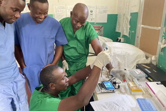 Lab staff from LFHV-GKD during hands-on sequencing training in loading and handling a MinION flow cell (Oxford Nanopore Technologies). November 2023, Gueckédou, Guinea. Drei Labormitarbeiter in professionellen blauen und grünen Kitteln beobachten einen Kollegen, der eine MinION flow cell befüllt (Oxford Nanopore Technologies).