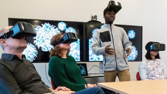 Several people wearing VR glasses. A man with his glasses pushed up talks to the group.