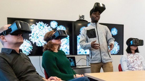 Mehrere Personen mit VR Brillen. Ein Mann spricht mit hochgeschobener Brille in die Runde