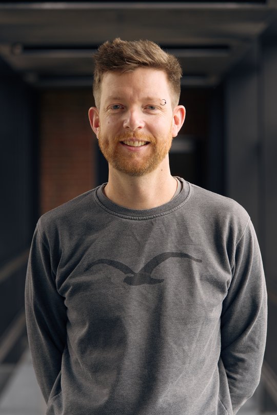 Dr. Benno Kreuels: ein Arzt mit kurzen rötlichen Haaren und einem kurzem rötlichen Bart in grauem Pullover. Über seiner linken Augenbraue trägt er einen Piercingring.