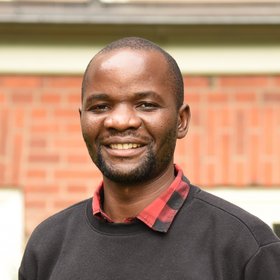 Das Bild zeigt einen Mann, der ein Hemd und einen Pullover trägt und freundlich in die Kamera schaut. Im Hintergrund sind die Backsteine eines Gebäudes zu sehen.