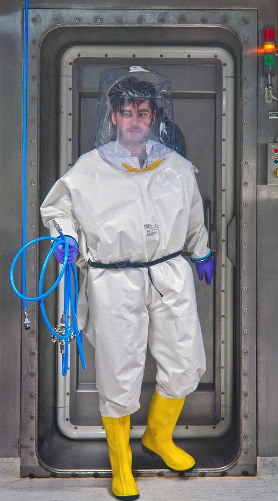 A researcher in a high-security laboratory wearing a full protective suit. A researcher leaves the airlock in the direction of the laboratory. He is wearing a white full suit and yellow rubber boots. He wears a transparent bonnet over his head. In his hand he carries a blue hose.