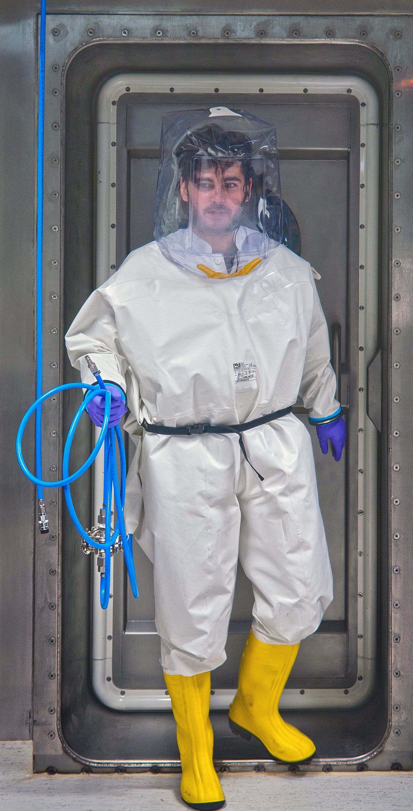 A researcher leaves the airlock in the direction of the laboratory. He is wearing a white full suit and yellow rubber boots. He wears a transparent bonnet over his head. In his hand he carries a blue hose.