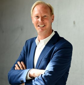 The photo shows a blond researcher dressed in a white shirt and dark blue jacket standing in front of a grey concrete wall. He smiles openly into the camera.