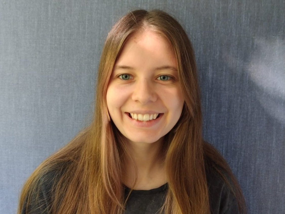 Eine junge Frau mit langen braunen Haaren schaut freundlich in die Kamera