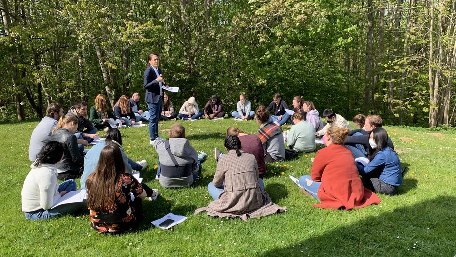 [Translate to English:] Eine Wissenschaftlerin steht auf dem Rasen im Kreis weiterer Teilnehmer:innen der LCI Summer School und hält einen Vortrag.