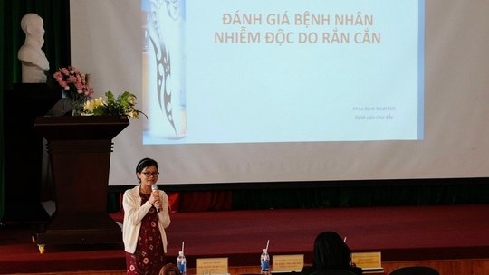 The picture shows a woman who is speaking into a microphone and standing in front of a projected presentation. On the presentation is Vietnamese writing.