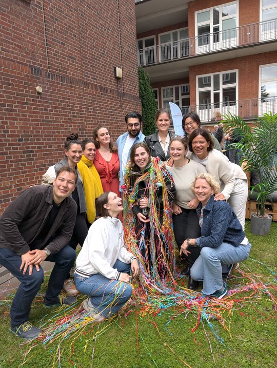 Barbara Honecker stands in the middle of her work group in the garden of the BNITM and is decorated with colourful streamers. Everyone in the picture is laughing happily into the camera