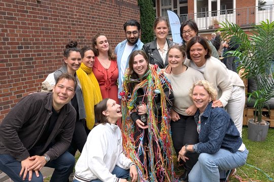 Barbara Honecker stands in the middle of her work group in the garden of the BNITM and is decorated with colourful streamers. Everyone in the picture is laughing happily into the camera