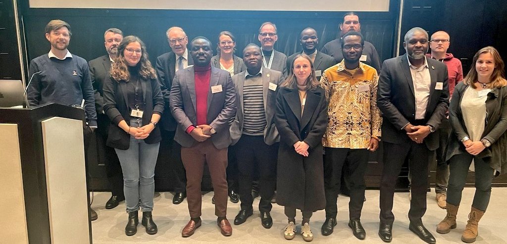 The picture shows a group of researchers from Europe and Africa standing in front of a screen and smiling friendly into the camera.