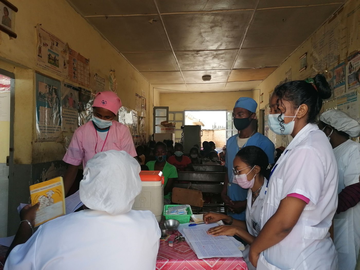 A group of nurses are standing around a small table and discussing documents that are lying on the table. Waiting patients can be seen in the background. All persons are wearing nose and mouth coverings.