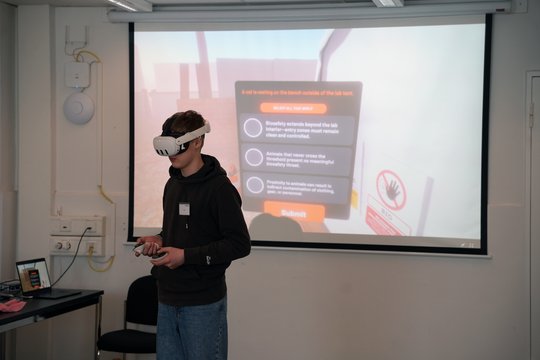 Auf dem Foto ist ein Schüler in dunklem Outfit zu sehen, der eine weiße VR-Brille trägt. Hinter ihm ist eine Präsentationsfolie mit einer Quizfrage auf einer Leinwand zu sehen.