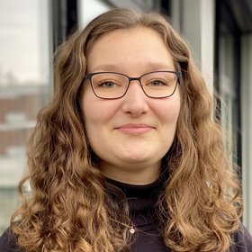 Das Bild zeigt eine Frau mit braunen lockigen Haaren, die ein schwarzes Oberteil trägt und freundlich in die Kamera schaut.