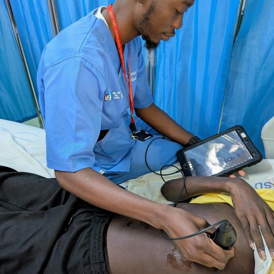 Das Bild zeigt einen Mann, der einen Patient mit einem Ultraschallgerät schallt. Währenddessen schaut er auf ein Tablet, auf dem der Ultraschall zu sehen ist.