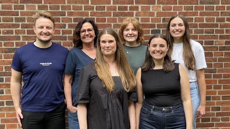 A group of six people smiles in front of a wall. They appear relaxed and happy and create a warm and friendly atmosphere.