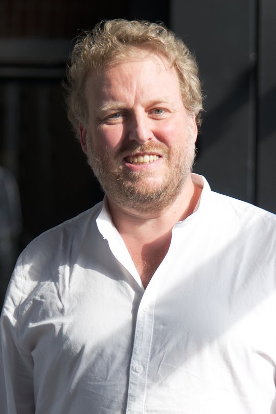 Dr. Florian Gehre Das Foto zeigt Dr. Florian Gehre. Er trägt kurze rötliche Locken, einen Dreitagebart und ein weißes Hemd und lächelt freundliche in die Kamera.