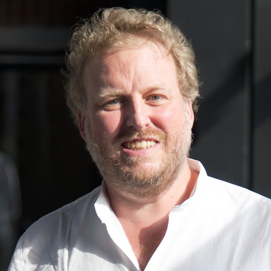 Dr. Florian Gehre Das Foto zeigt Dr. Florian Gehre. Er trägt kurze rötliche Locken, einen Dreitagebart und ein weißes Hemd und lächelt freundliche in die Kamera.