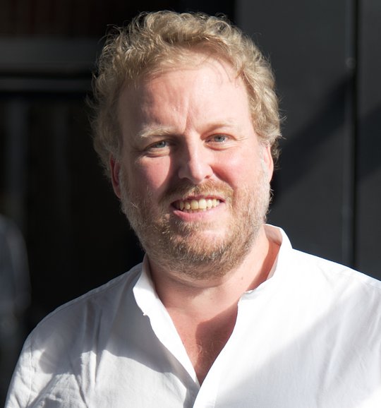 Dr. Florian Gehre Das Foto zeigt Dr. Florian Gehre. Er trägt kurze rötliche Locken, einen Dreitagebart und ein weißes Hemd und lächelt freundliche in die Kamera.
