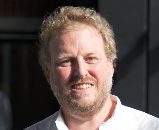 Das Foto zeigt Dr. Florian Gehre. Er trägt kurze rötliche Locken, einen Dreitagebart und ein weißes Hemd und lächelt freundliche in die Kamera.