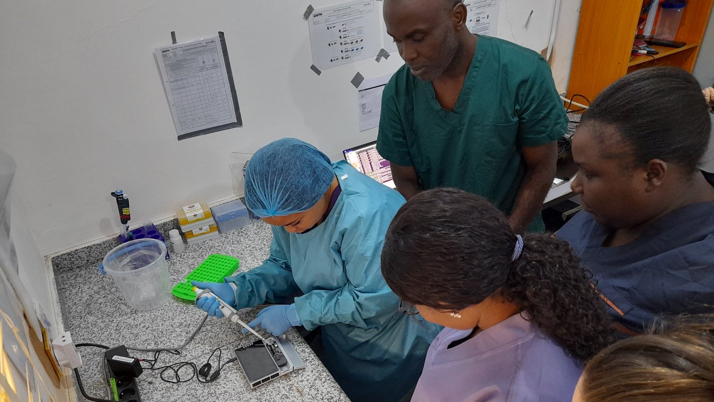 ISTH sequencing lab staff loading sequencing samples in a MinION flow cell The photo shows a Nigerian lab team of four and their trainer at a lab bench. They wear blue and green lab coats and gloves. One of them is loading a sequencing device with a pipette while the others observe.
