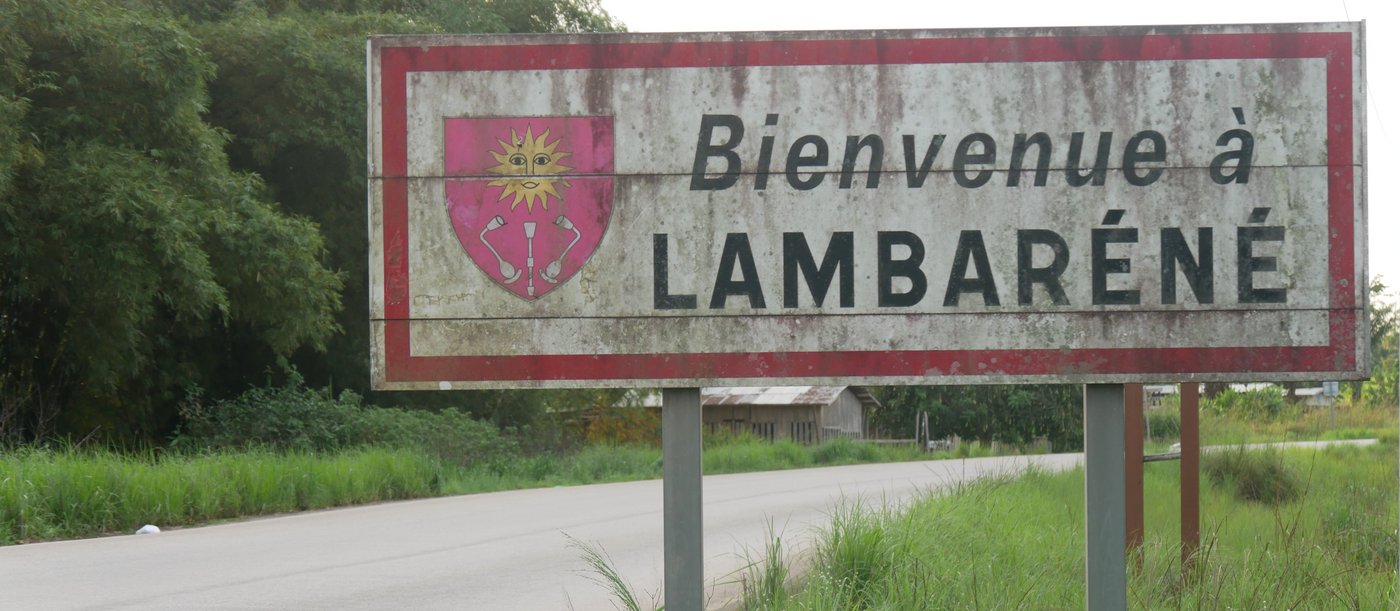 The picture shows a road with green grass and trees at the roadside. On the right side of the road is a white sign with a red edge. On the sign is a red coat of arms and the inscription "Bienvenue à Lambaréné".