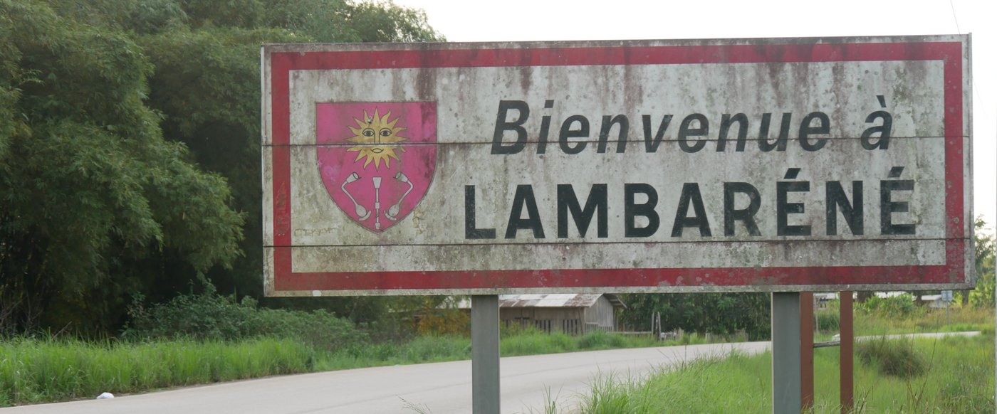Das Bild zeigt eine Straße mit grünem Gras und Bäumen am Straßenrand. Auf der rechten Seite der Straße steht ein weißes Schild mit rotem Rand. Auf dem Schild befindet sich ein rotes Wappen und die Aufschrift "Bienvenue à Lambaréné".