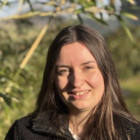 Birte Viebrock A person with long hair smiles outdoors in a sunlit setting with green foliage and bamboo in the background.