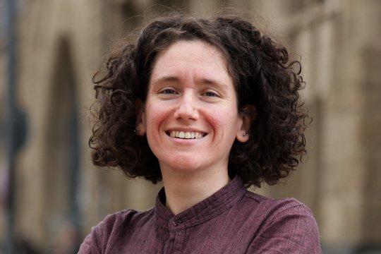 Dr. Giuditta Annibaldis: eine lächelnde Forscherin mit dunklen Locken vor einer hellbraunen Häuserfassade. Sie trägt ein weinrotes Hemd und verschränkt die Arme vor dem Körper.